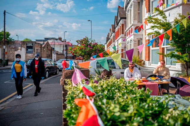 People wearing protective face masks walk past Walthamstow residents having a street party in their front garden to celebrate the 75th anniversary of VE Day (Victory in Europe Day), the end of the Second World War in Europe in Walthamstow in East London on May 8, 2020. (Photo by Tolga AKMEN / AFP) (Photo by TOLGA AKMEN/AFP via Getty Images)