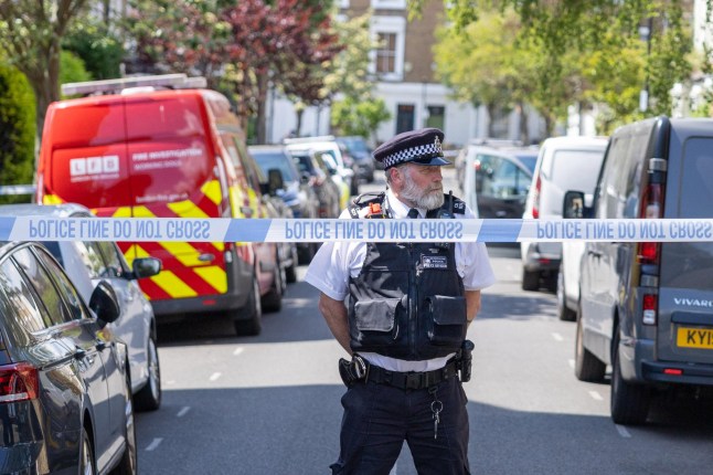 A police officer standing in a London street, behind a 'police line do not cross' banner.