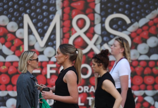 Shoppers pass in front of an M&S logo on a wall.
