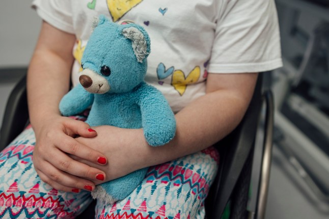 A close-up shot of an unrecognisable young girl sitting in a wheelchair embracing a teddy bear whilst on a routine visit to hospital in Newcastle upon Tyne, North East England. The young girl is wearing colourful casual clothing and has her hands clasped in front of the teddy bear, which she has brought along with her to provide comfort.