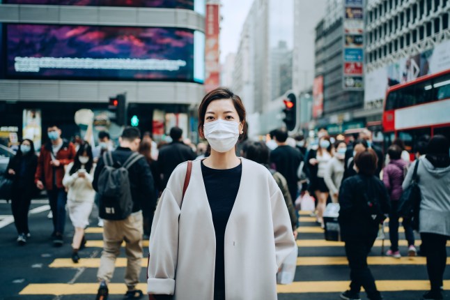 Portrait of young Asian woman with protective face mask standing in the middle of busy downtown city street amidst crowd of pedestrians passing by, looking at the camera, with highrise city buildings and busy traffic in background (Credits: Getty Images)