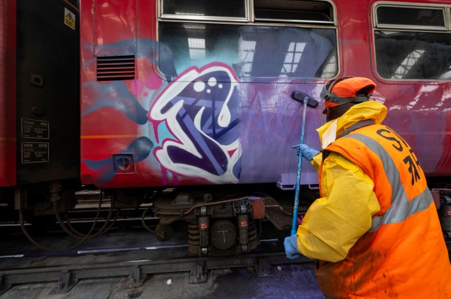 A train presentation staff member cleaning a carriage off graffiti inside a depot building with a brush.