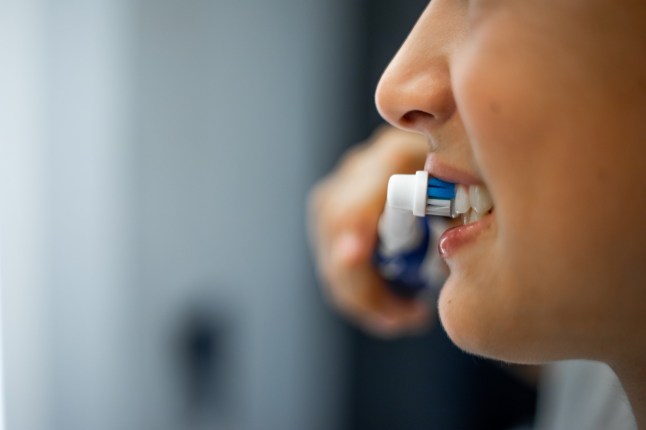 Close up mouth of teenage boy holding electric toothbrush in hand, smiling and brushing white teeth with modern equipment. Loveable teenager performing everyday procedure, caring of dental health in bathroom.