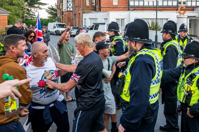 BNPS.co.uk (01202 558833) Pic: MaxWillcock/BNPS Pictured: The protest outside the hotel. This is the moment an asylum seeker gives protesters the finger as he enters a migrant hotel. He was being escorted by a police officer into The Chine Hotel in Bournemouth, Dorset, when 40 protesters stood outside the premises started shouting abuse at him. The migrant stops in his tracks and turns around to give the man the unsavoury hand gesture before a second police officer gets involved as tensions flare. The late 19th century hotel on the seafront was once a favourite of celebrities including Stan Laurel, Oliver Hardy and Vera Lynn.
