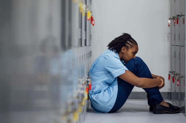 Medium shot of a female nurse sitting down in the locker room. She is hunched forward with her arms wrapped around her legs. Her back is against the lockers and she is looking away from the camera. The woman is wearing a blue nurse uniform.