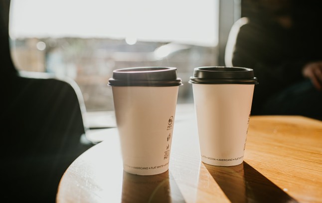 Two backlit disposable takeaway cardboard coffee cups with plastic lids sit on a wooden surface casting long shadows. Space for copy.