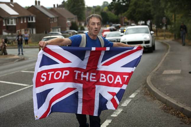 LONG EATON, ENGLAND - AUGUST 23: A protester against the hotel holds a Union flag with 'Stop The Boats' written on it outside the Best Western hotel during the demonstration on August 23, 2025 in Long Eaton, United Kingdom. Far-right protests at migrant hotels across the UK have been a feature of the summer months since demonstrations at The Bell Hotel in Epping earlier this summer. (Photo by Martin Pope/Getty Images)