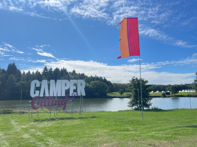 Ragley Hall-Alcester-UK-August 23,2023 : A large brightly coloured flag lit by the sun on a blue sky summer day with Camper Calling sign at the music festival.Lake and tents visible in background; Shutterstock ID 2351418709; purchase_order: -; job: -; client: -; other: -