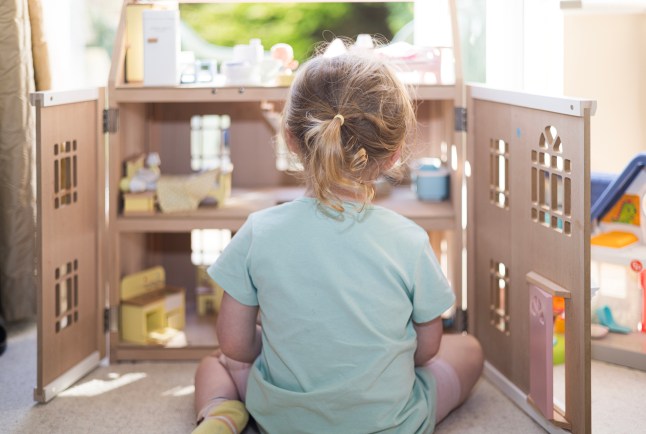 Lonely three year old girl playing on her own with her wooden dolls house in a domestic living room
